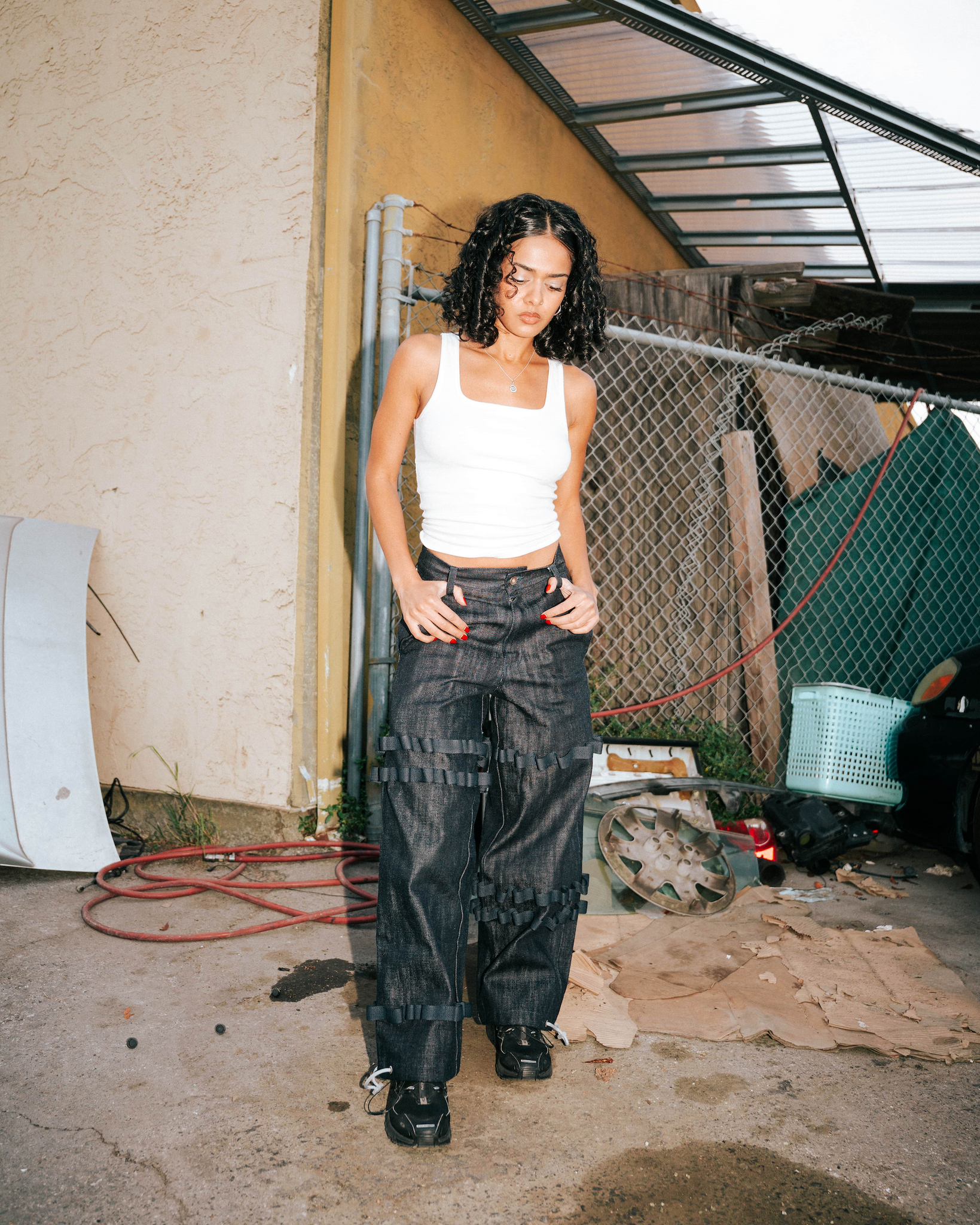 Person wearing a white tank top and black pants standing in an outdoor setting with a fence and various items in the background.